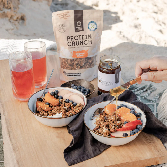 Two bowls of granola with fruit, a jar of honey, and a bag of protein crunch on a wooden table.
