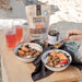 Two bowls of granola with fruit, a jar of honey, and a bag of protein crunch on a wooden table.